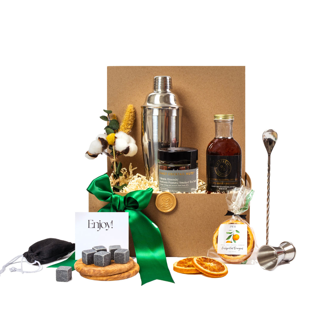 A luxury sustainable gift basket with
a cocktail shaker, cocktail syrup, dried oranges, cocktail spoon, wooden coasters, whiskey stones and premium cocktail nuts