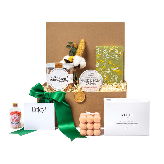 Gift basket with matches, shortbread cookies, artisanal cube candle, hand and body cream, mini notebook, Sitti natural body sponges finished with dried seasonal greenery.