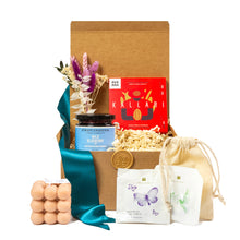 Eco-friendly gift basket featuring sustainable products, including wild blueberry jam, artisan chocolate, herbal teas, a natural soy wax candle, dried flower bouquet, and a reusable cotton pouch, all presented in a cardboard box with teal ribbon. Perfect for housewarming or corporate gifting