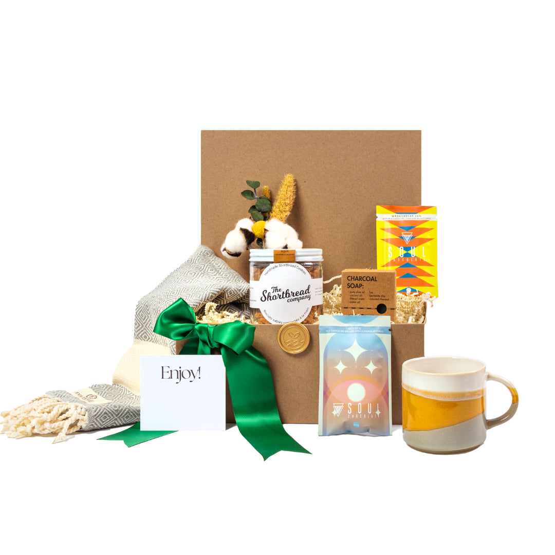 A kraft gift basket with soul chocolate hot chocolate, ceramic mug, cotton throw, shortbread cookies and charcoal soap. 