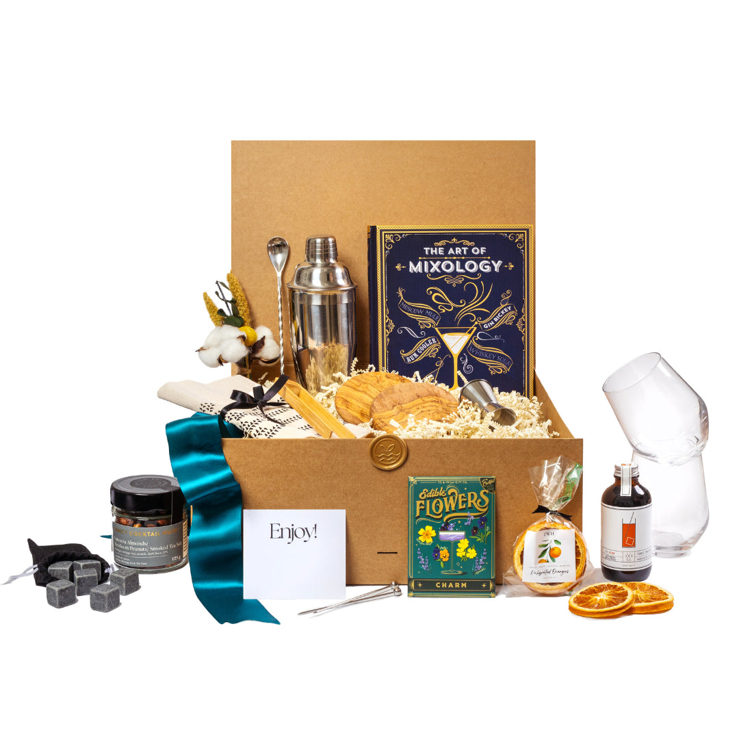 A cocktail gift basket with a mixology book, cocktail nuts, edible flowers, cocktail shaker, cocktail spoon, organic napkin, seasonal greenery, cocktail syrup